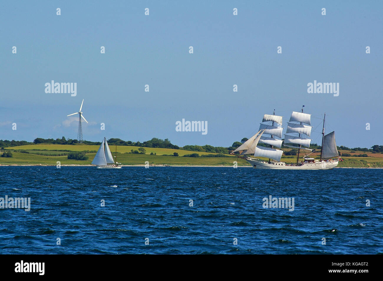 Piazza tradizionale-truccate tall ship e moderna a due-masted yacht in mare davanti a un mare verde con campi e wind power station Foto Stock