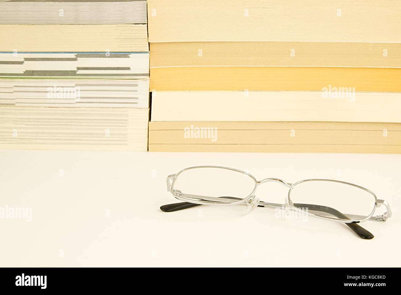 Pila di libri romanzi su un bianco superficie riflettente con alcuni bicchieri d'argento (occhiali) giacenti in primo piano. Lettura del concetto di alfabetizzazione Foto Stock