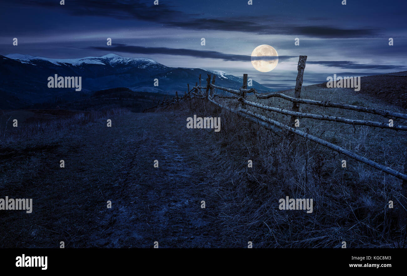 Recinzione in legno in campagna montuosa di notte in luna piena luce. crinale montuoso con cime innevate della distanza Foto Stock
