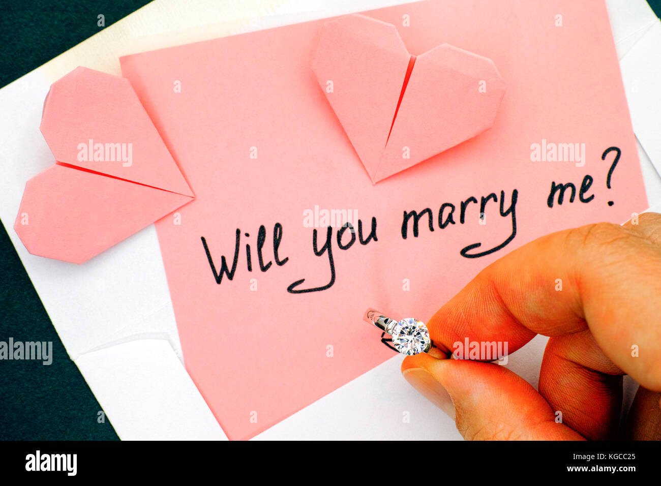 Donna mano azienda anello di fidanzamento. Lettera con testo ti sposerà me? In busta e due cuori origami sul tavolo. Foto Stock