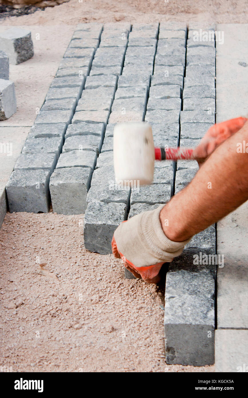 Le mani nei guanti di un lavoratore builder posa sul granito imp ciottoli sulla sabbia con un mazzuolo di gomma martello , e la pavimentazione stradale sul marciapiede costrut Foto Stock