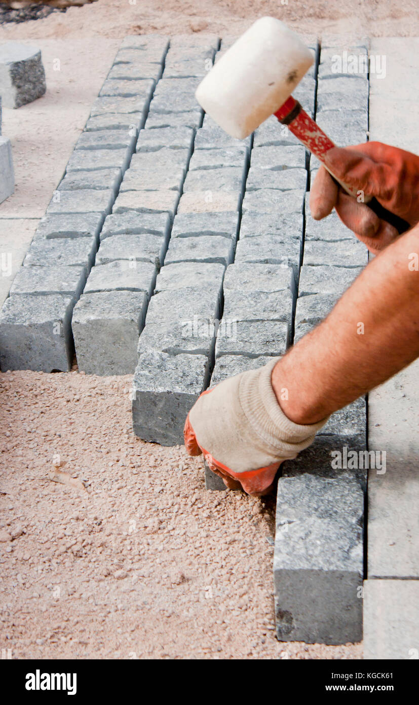 Le mani nei guanti di un lavoratore builder posa sul granito imp ciottoli sulla sabbia con un mazzuolo di gomma martello , e la pavimentazione stradale sul marciapiede costrut Foto Stock