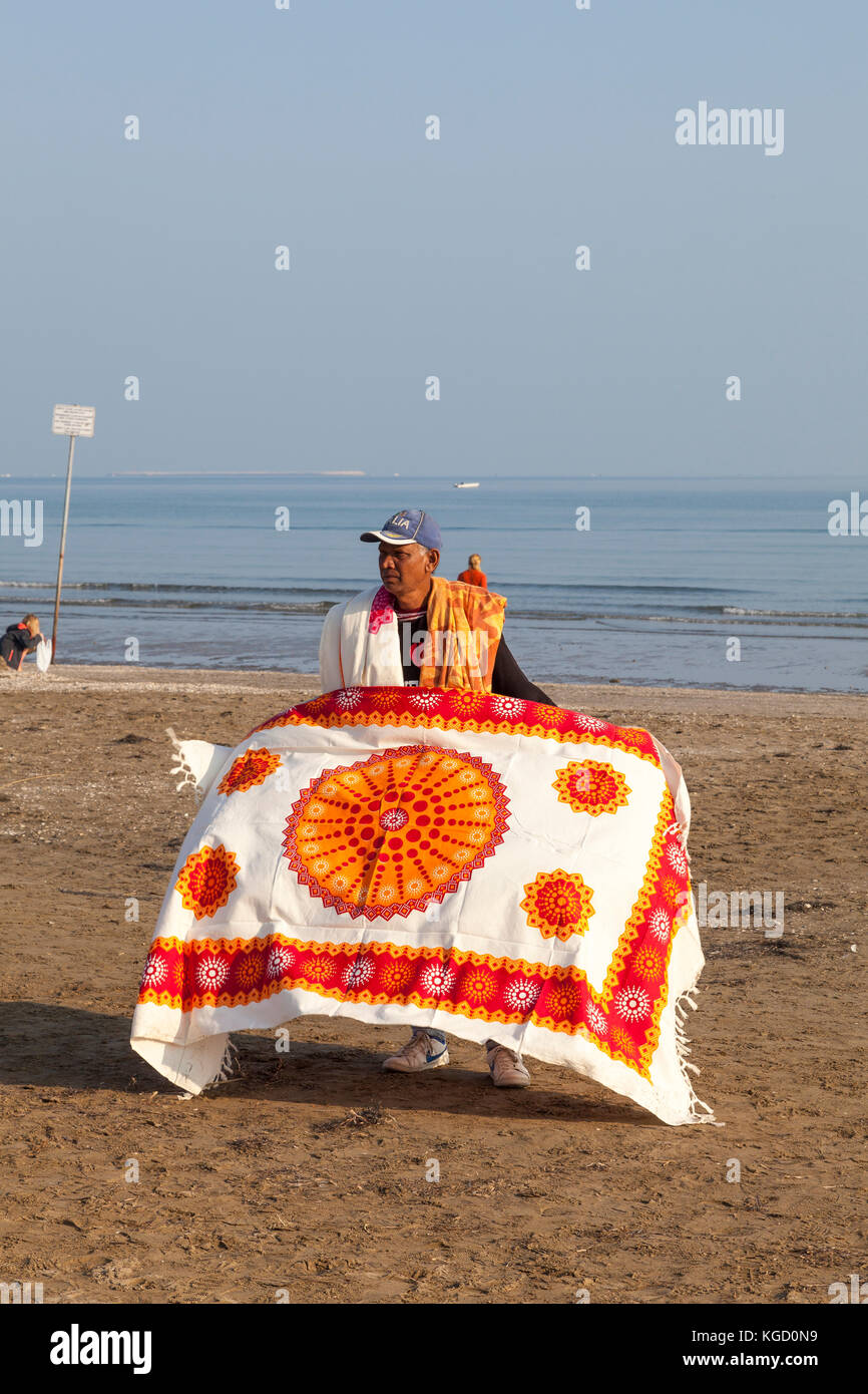 Fornitore di migranti visualizzando un vivacemente colorato tappeto in vendita Blue Moon Beach, Lido di Venezia al tramonto come egli cerca di attirare i turisti Foto Stock