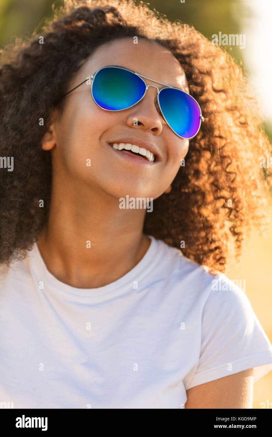 Una bella e felice di razza mista americano africano ragazza adolescente figlio giovane donna nella luce del sole che indossa blu riflettente aviator occhiali da sole e sm Foto Stock