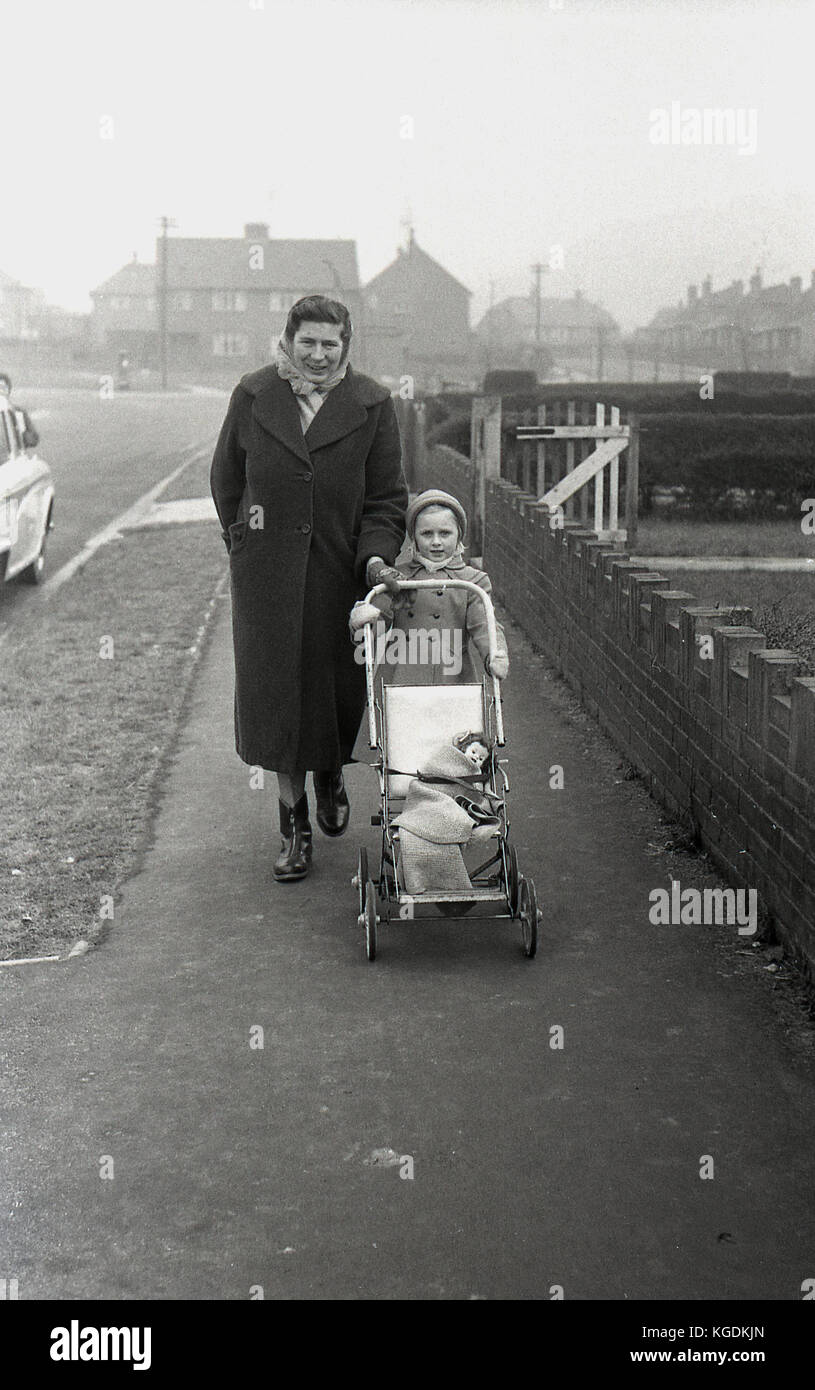 Degli anni Cinquanta, storico, madre avvolto nel mantello e sciarpa a spasso con la sua giovane daugher che sta spingendo un piccolo pram o buggy dell'epoca con la sua bambolina che è avvolto con una coperta, Inghilterra, Regno Unito. Foto Stock