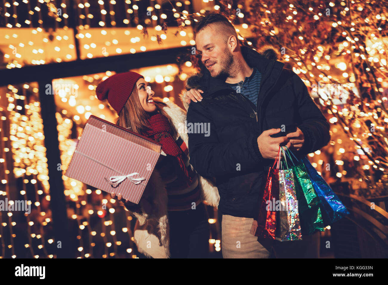 Coppia giovane in piedi al di fuori con le borse della spesa e scatole e godendo di una passeggiata serale attraverso la città al tempo di Natale. Foto Stock