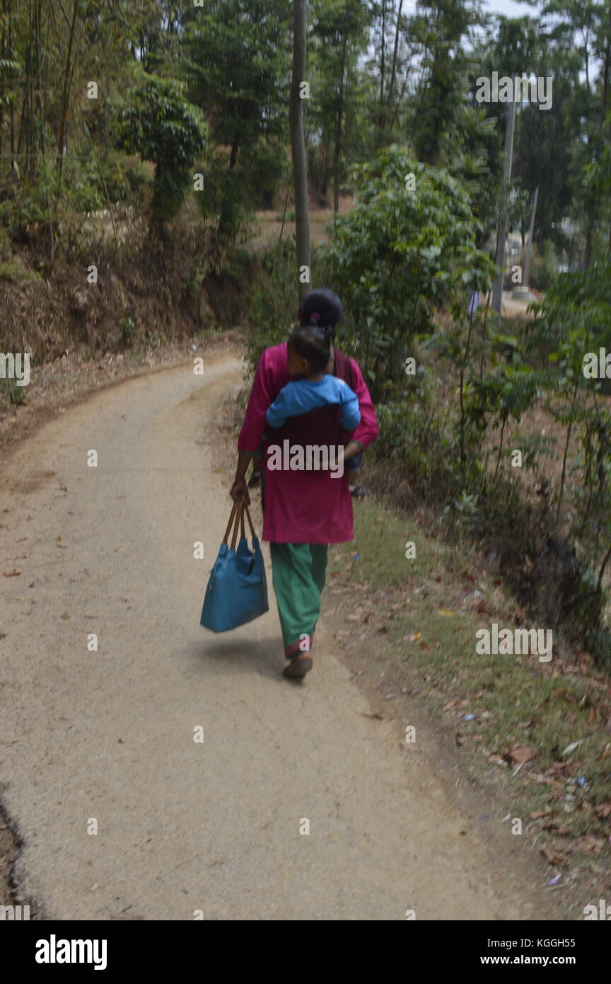 Donna nepalese con il bambino sulla schiena camminando su una strada sterrata in un piccolo villaggio, Panauti, nelle montagne himalaya del Nepal. Kameez tradizionale. Foto Stock