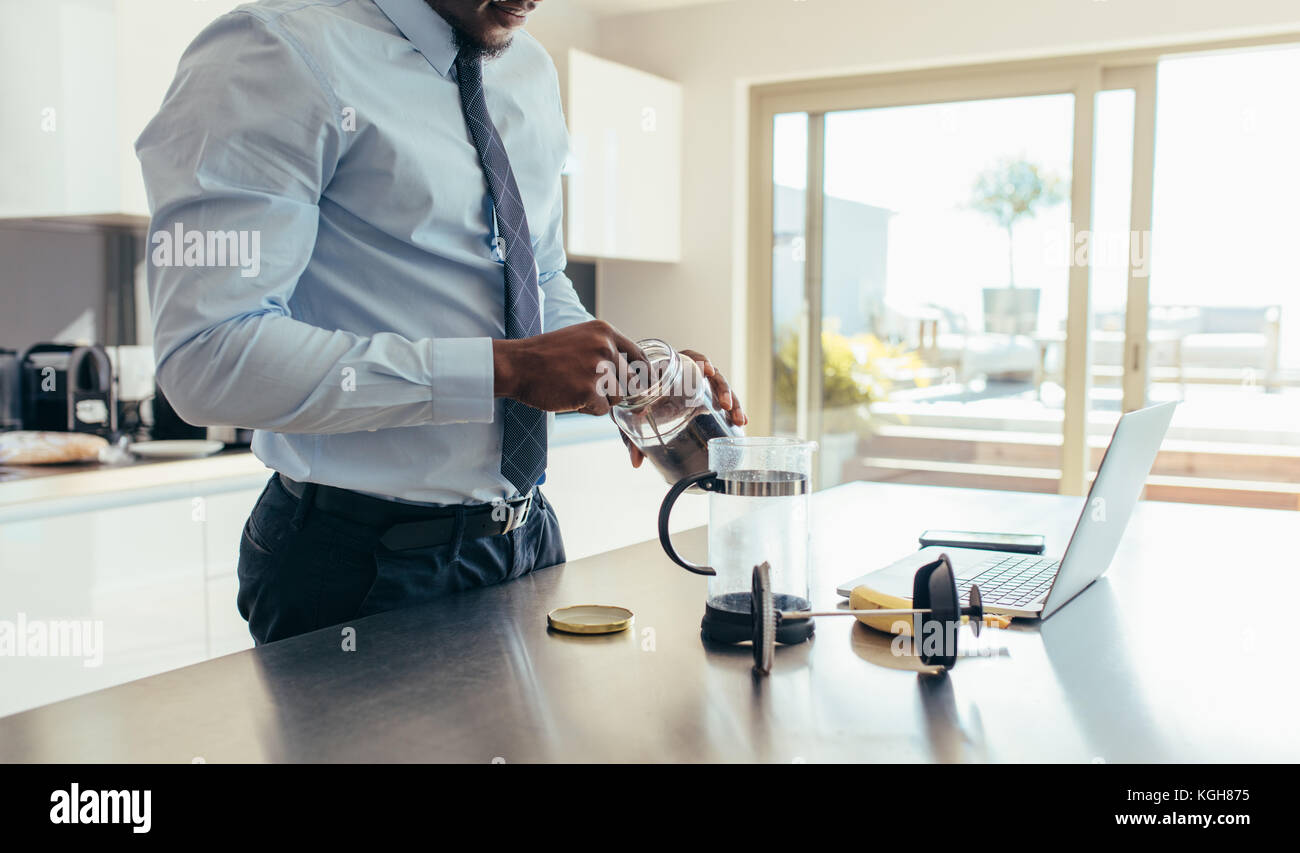 Uomo in abbigliamento formale il caffè utilizzando una French press con un computer portatile sul tavolo da cucina. Imprenditore preparazione di caffè mentre facendo il lavoro di ufficio di un Foto Stock