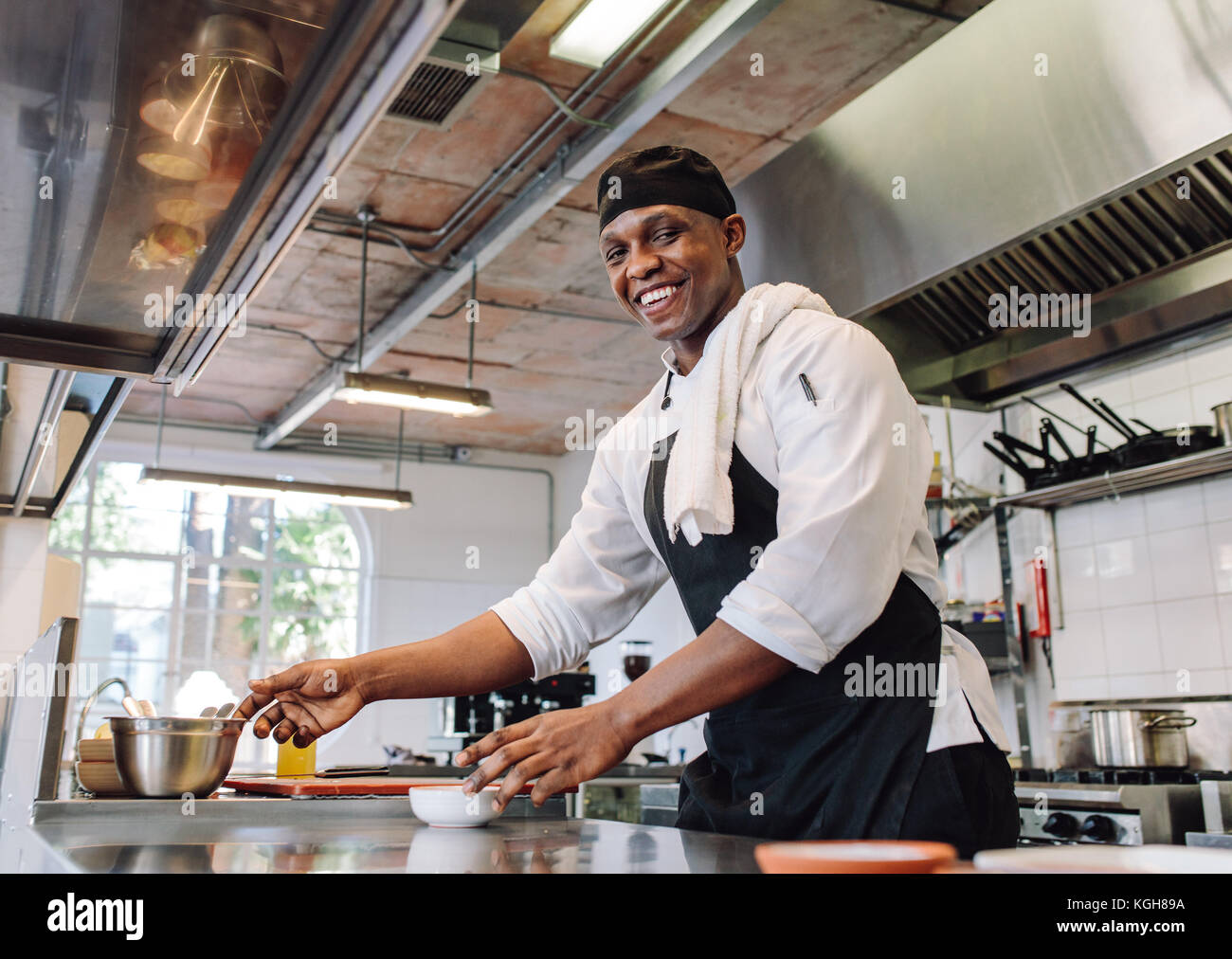 Lo chef sorridente la cottura di cibo al ristorante Cucina. Maschio africano cuocere in piedi dal bancone cucina guardando la fotocamera e sorridente. Foto Stock