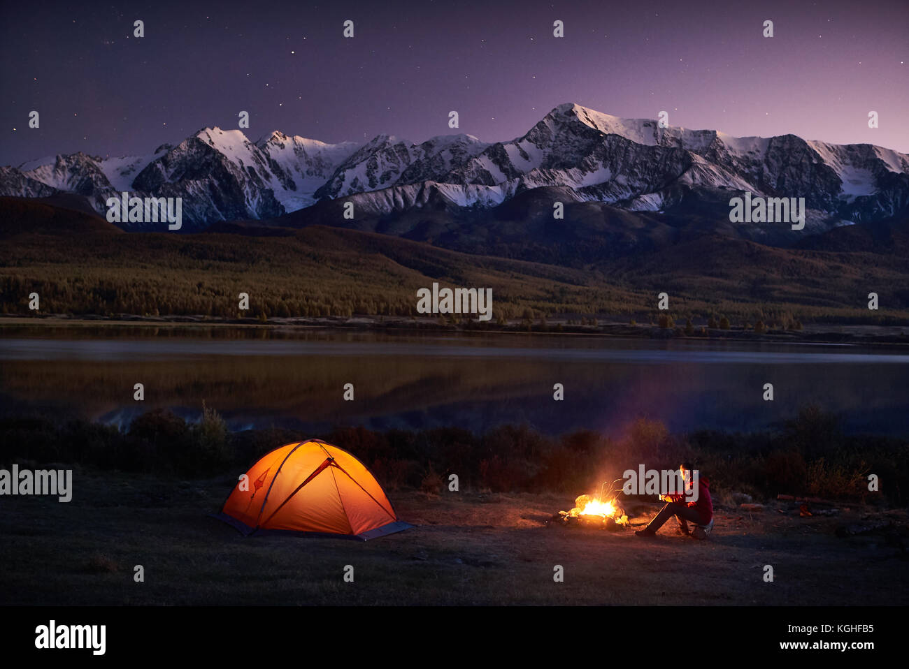 Notte Campeggio. turisti uomo seduto nella tenda illuminata vicino al falò sotto un tramonto meraviglioso cielo di sera in una zona di montagna. neve montagna in b Foto Stock