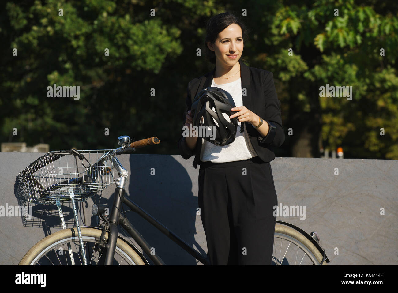 Sorridente imprenditrice tenendo il casco in piedi in bicicletta contro piante sulla giornata di sole Foto Stock