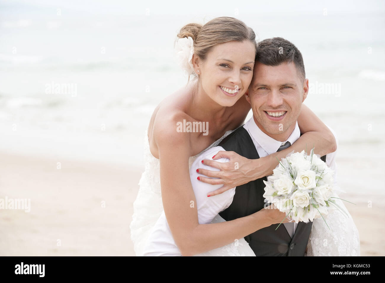 Lo sposo dando piggyback ride alla sposa sulla spiaggia Foto Stock