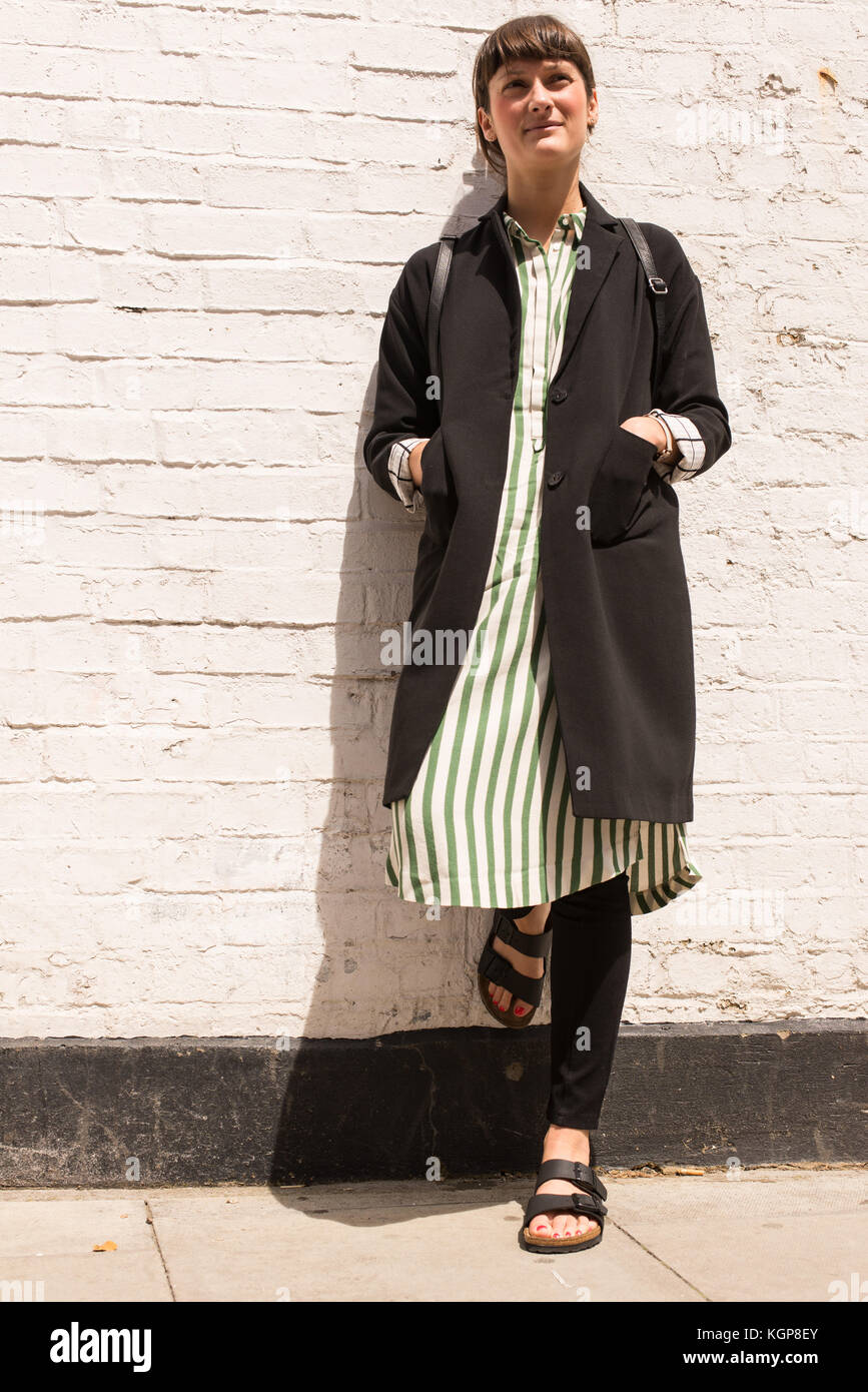 Hipster donna vestita di urbano in stile minimale con un lungo mantello nero a strisce abito, pantaloni neri e sandali. shot su una strada con il bianco muro di mattoni Foto Stock