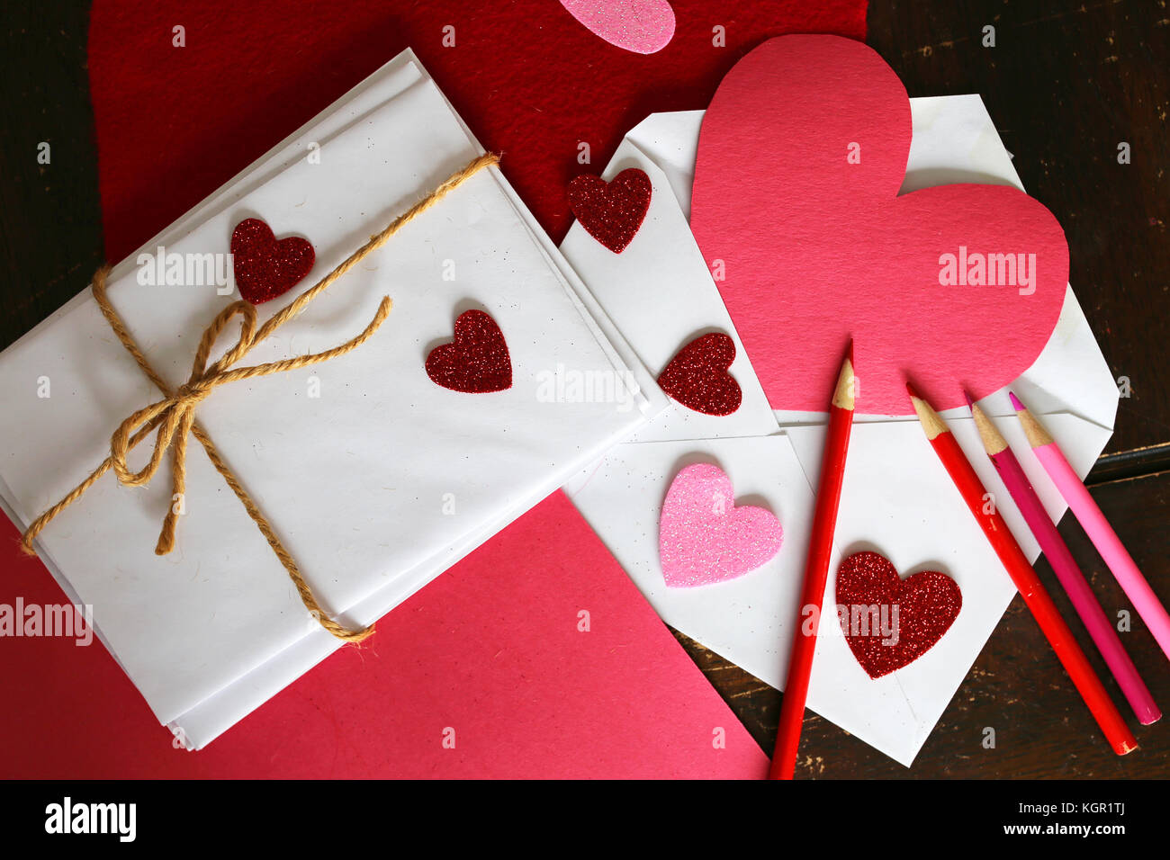 Il giorno di San Valentino lettera forniture d'arte tra cui: rosso e rosa matite colorate, costruzione della carta, a forma di cuore ad adesivi, e una pila di buste con lembi di chiusura Foto Stock