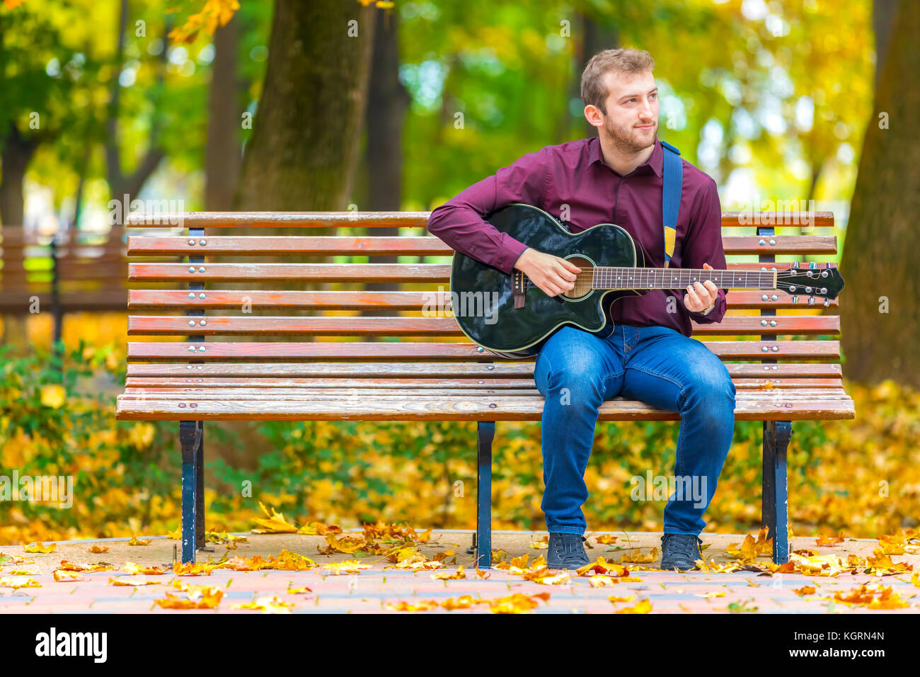 Giovane uomo seduto su un banco da lavoro e suonare la chitarra acustica nel parco della città Foto Stock
