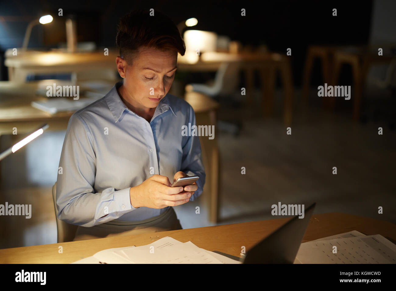 Occupato ragioniere texting nel suo smartphone durante la pianificazione di rimanere in ufficio per la notte Foto Stock