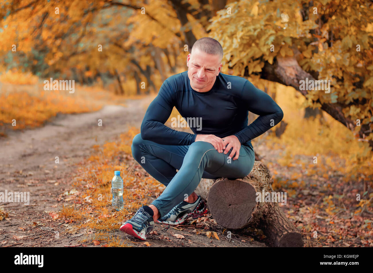 Un giovane pareggiatore di atletica in nero aderente sportswears e sneakers siede su un registro, si sente un forte dolore nel muscolo dopo crampi su un autumna Foto Stock