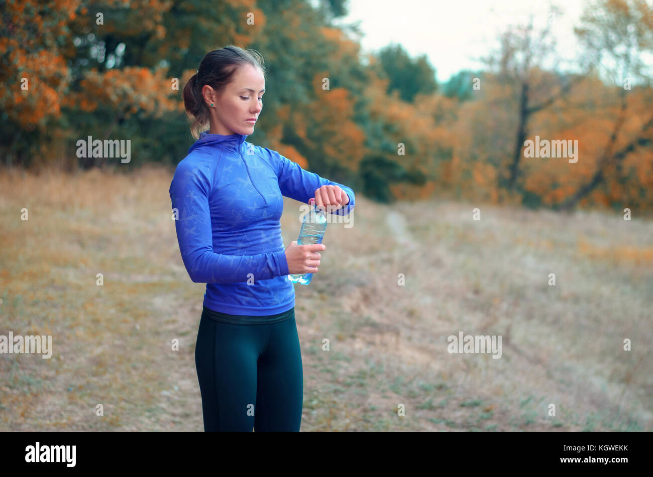 Un giovane caucasico donna atletica in un blu giacca sportiva con una cappa e leggins nero apre una bottiglia con acqua dopo il jogging su una foresta autumnall Foto Stock