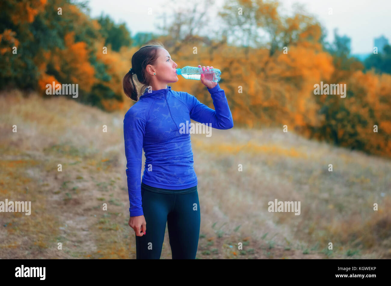 Un sottile pareggiatore caucasica donna in blu giacca sportiva con una cappa e leggins nero bere acqua dalla bottiglia dopo il jogging su un luminoso autumnall f Foto Stock