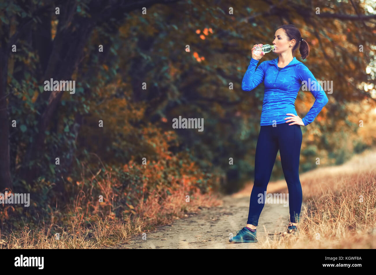 Un giovane pareggiatore atletica donna in blu giacca sportiva con una cappa e leggins nero bere acqua dalla bottiglia dopo il jogging in un colorato autumnall Foto Stock