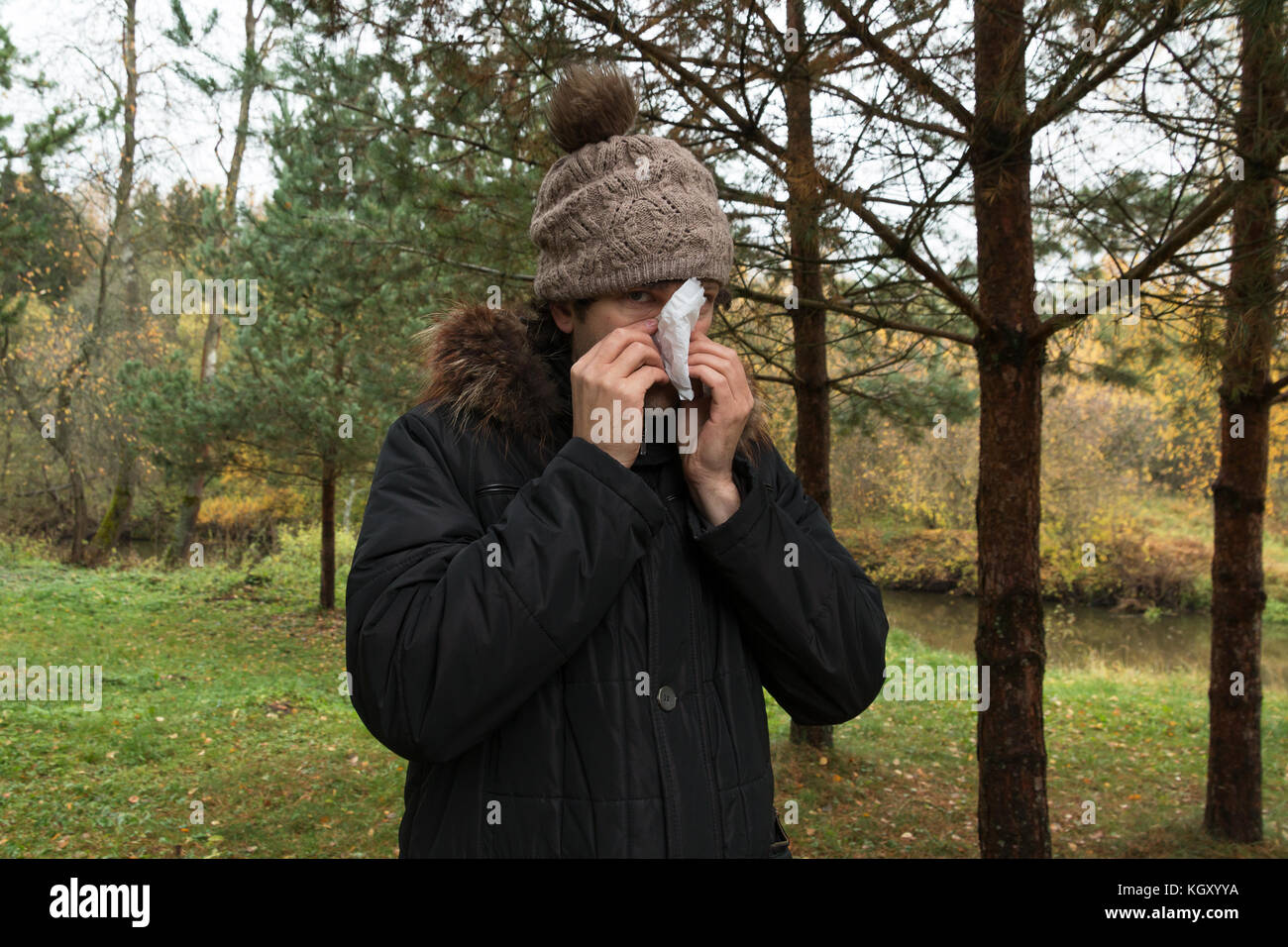 Giovane uomo in giacca soffrono di freddo e tenendo un fazzoletto sul naso. maschio in berretto lavorato a maglia in piedi in legno soffia il naso. autunno illne Foto Stock