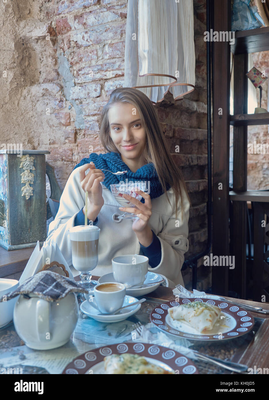 Un affascinante giovane ragazza dai capelli scuri è a riposo in un caffè accogliente. Il caffè è sul suo tavolo. La ragazza gode il gusto di gelato. Foto Stock