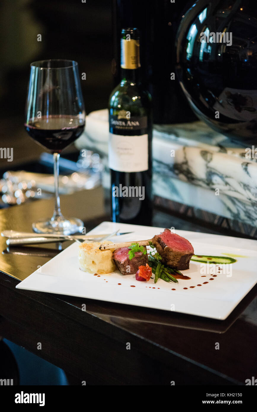 Un piatto di agnello accompagnato da una bottiglia e un bicchiere di vino rosso in cima a un tavolo nero Foto Stock
