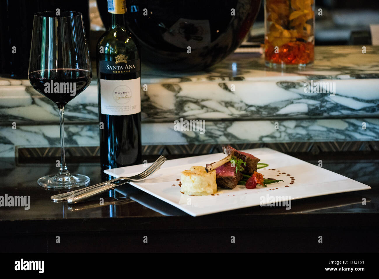Un piatto di agnello accompagnato da una bottiglia e un bicchiere di vino rosso in cima a un tavolo nero Foto Stock