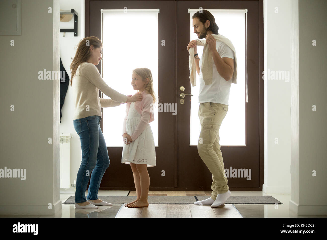 Madre premurosa aiutando medicazione piccola figlia per camminare con padre, famiglia felice in piedi a casa porta di ingresso, amorevole mamma e papà di trascorrere del tempo tak Foto Stock