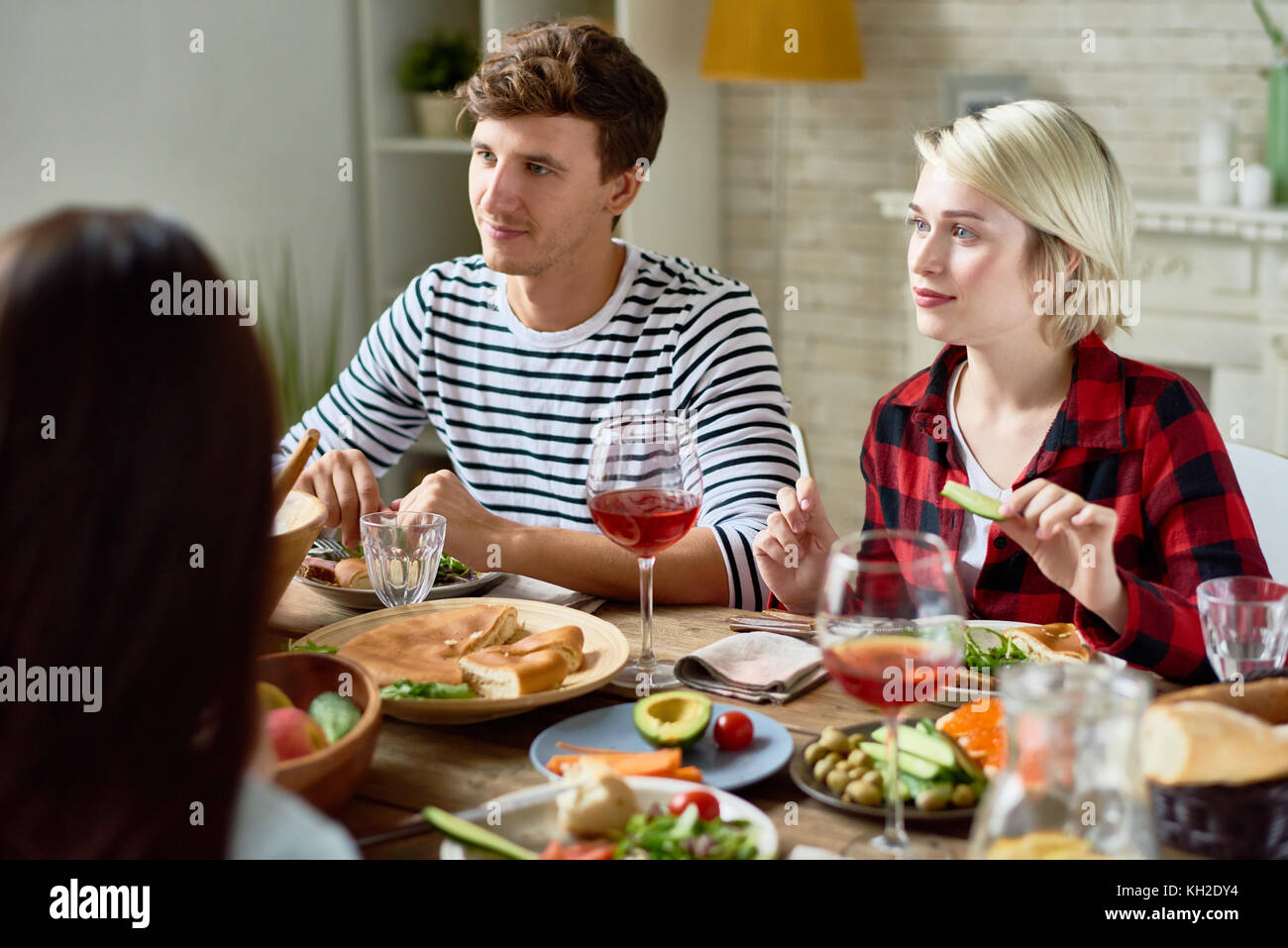 Ritratto di gruppo di giovani amici a cena insieme, riuniti al grande tavolo con deliziosi piatti su di esso, concentrarsi sul giovane ascoltando la conversazione Foto Stock