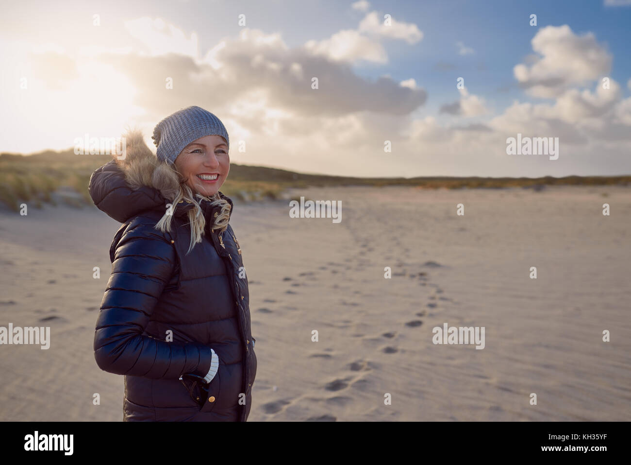 Moda donna in un caldo autunno vestito in piedi su una spiaggia al tramonto la colata di una lunga ombra attraverso la sabbia guardando al lato con un sorriso Foto Stock