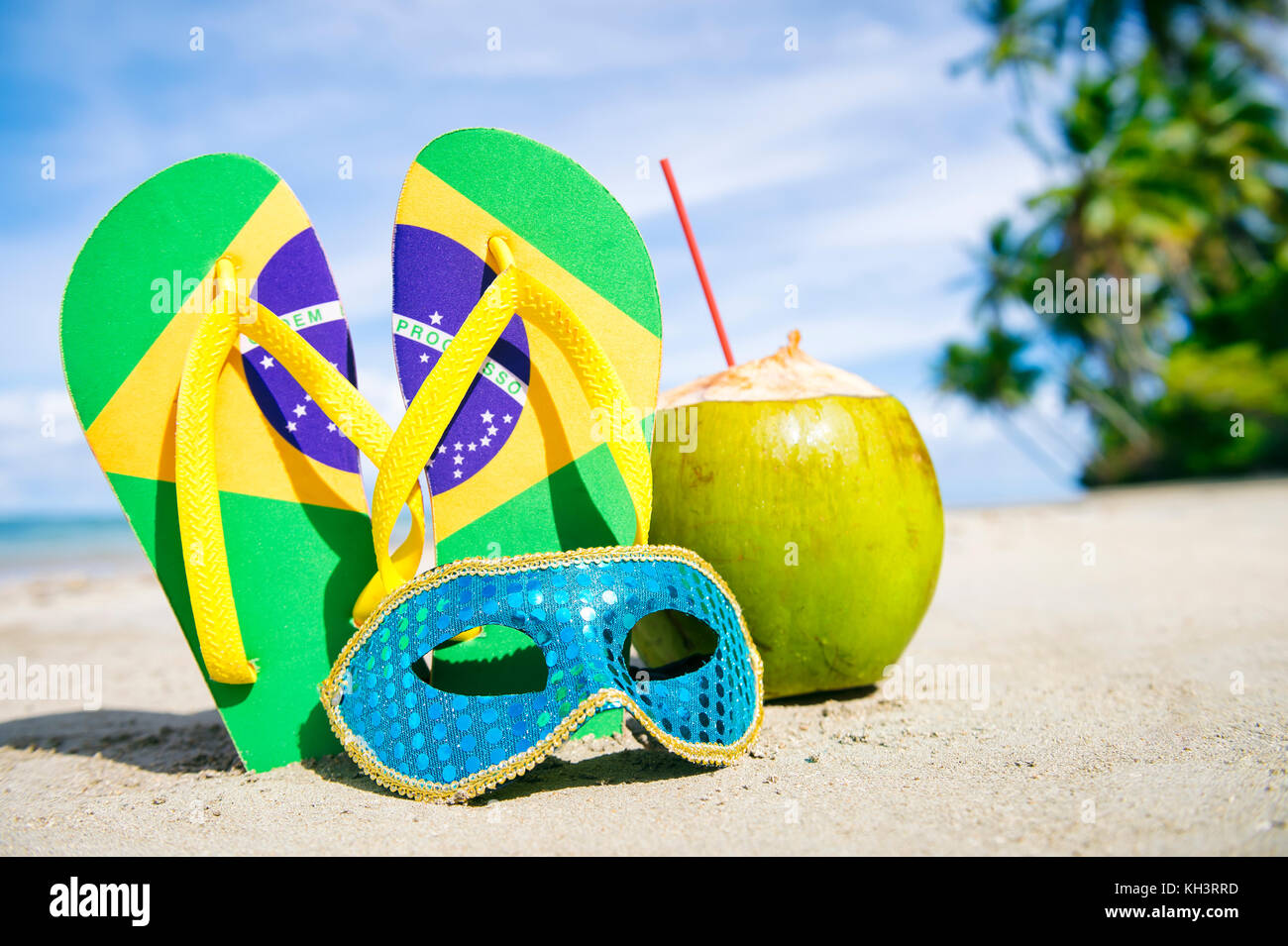 Bandiera Brasile flip flop, colorato sequined maschera di Carnevale e verde fresco drink di cocco su un tropicale spiaggia orlata di palme. Foto Stock