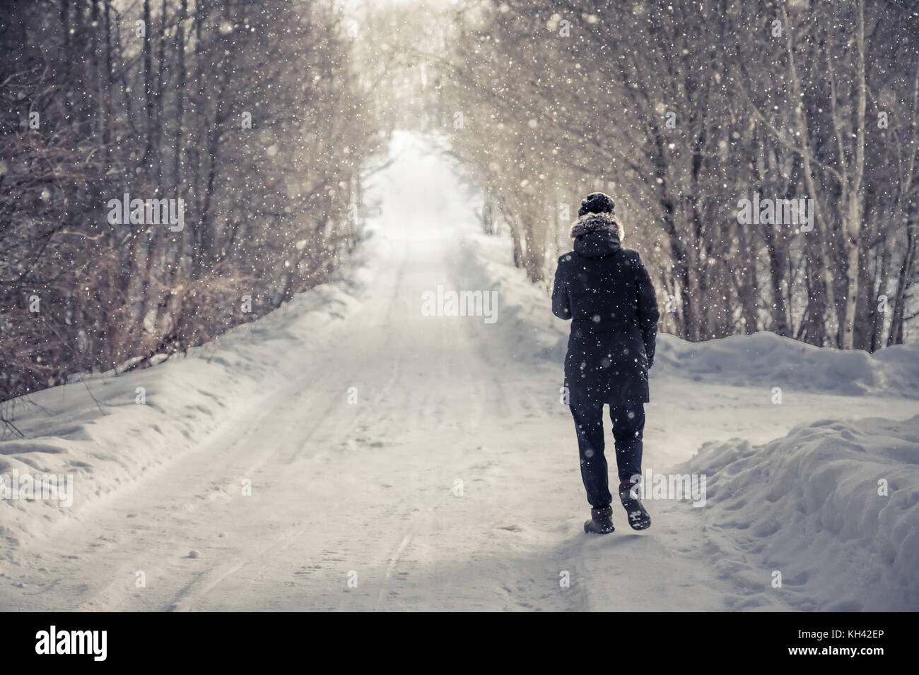 Donna solitarie passeggiate sulla neve in inverno road tra alberi alley con luce alla fine della strada in inverno freddo giorno durante la nevicata con spazio di copia Foto Stock