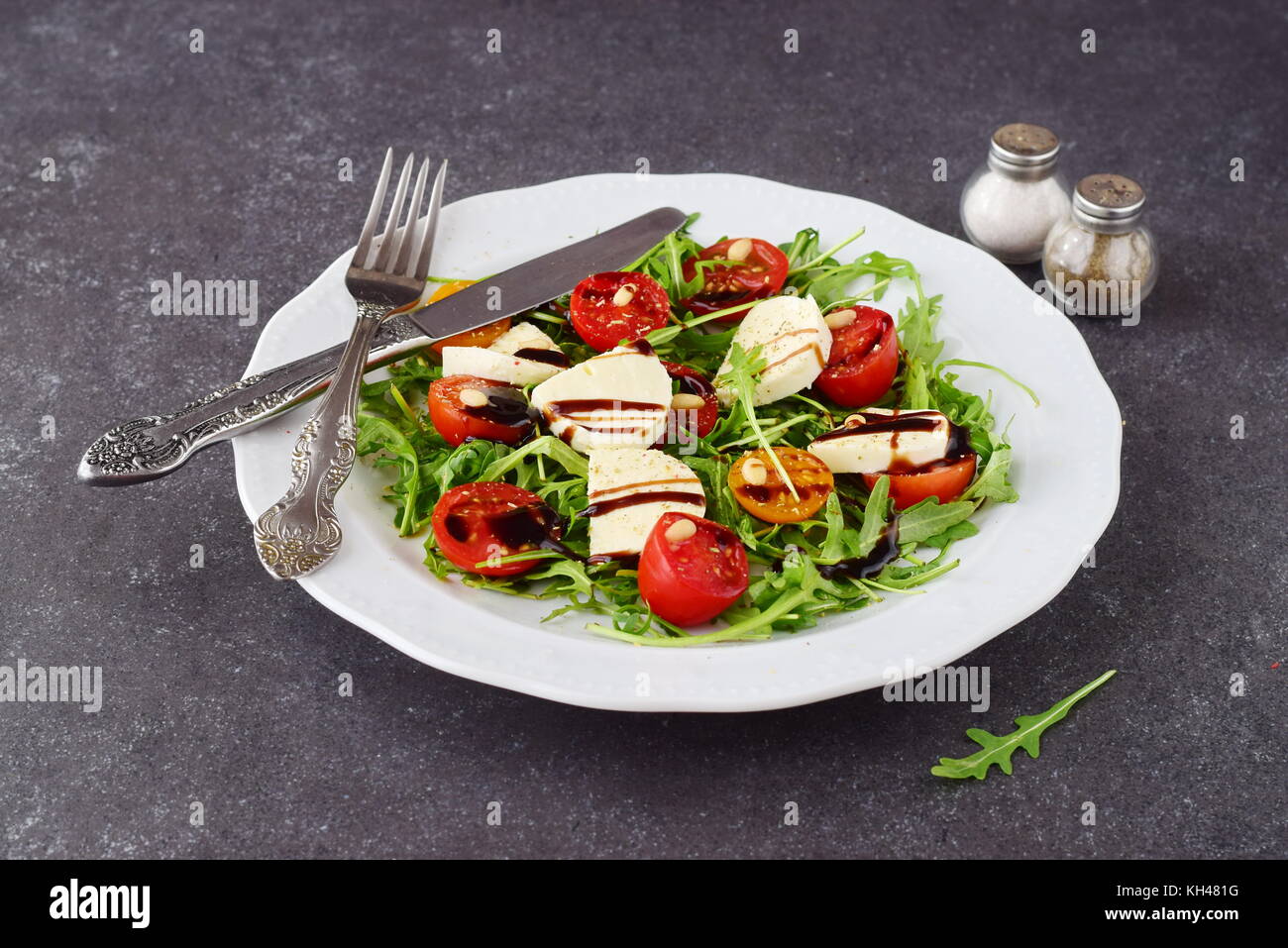 Tradizionale italiano con insalata di pomodoro ciliegino, rucola, mozzarella, olio di oliva aceto di vino su una piastra bianca su un grigio Sfondo astratto. Foto Stock