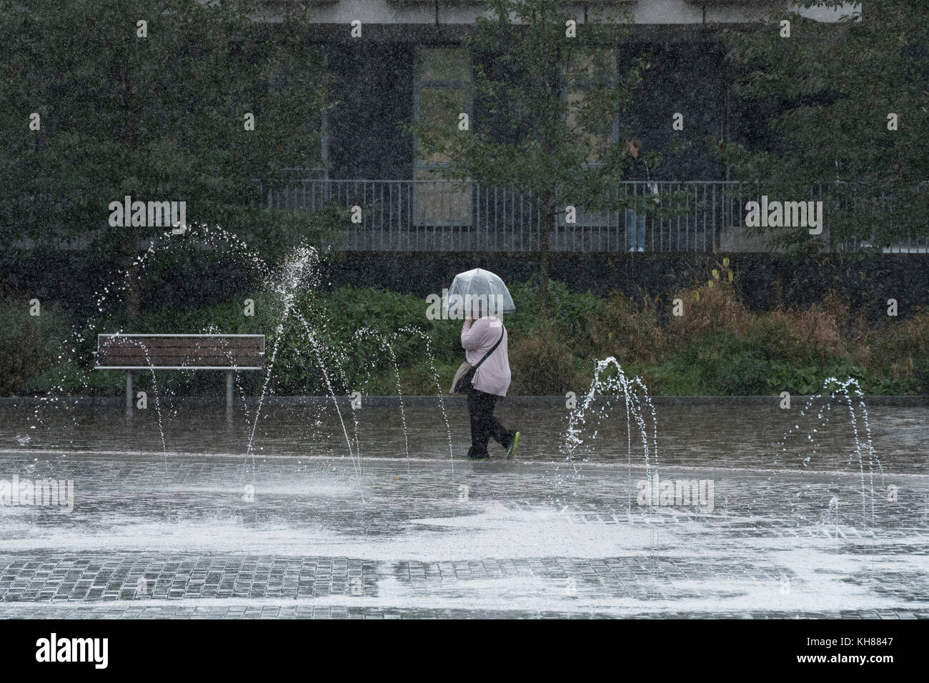 Donna che cammina in heavy rain, rifugiandosi sotto ombrellone, passando la piscina a specchio e fontane - Bradford City Park, West Yorkshire, Inghilterra, Regno Unito. Foto Stock