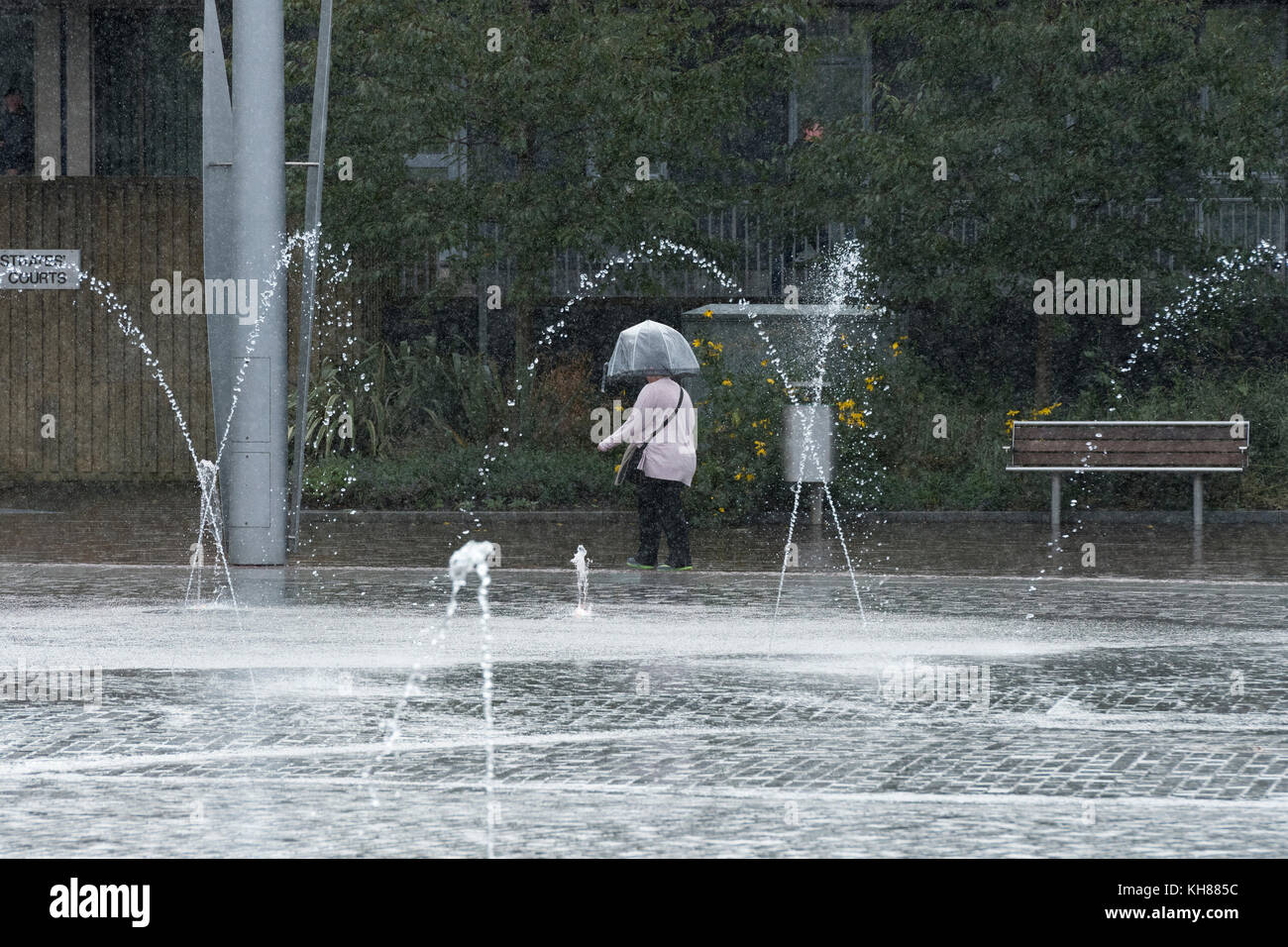 Donna che cammina in heavy rain, rifugiandosi sotto ombrellone, passando la piscina a specchio e fontane - Bradford City Park, West Yorkshire, Inghilterra, Regno Unito. Foto Stock