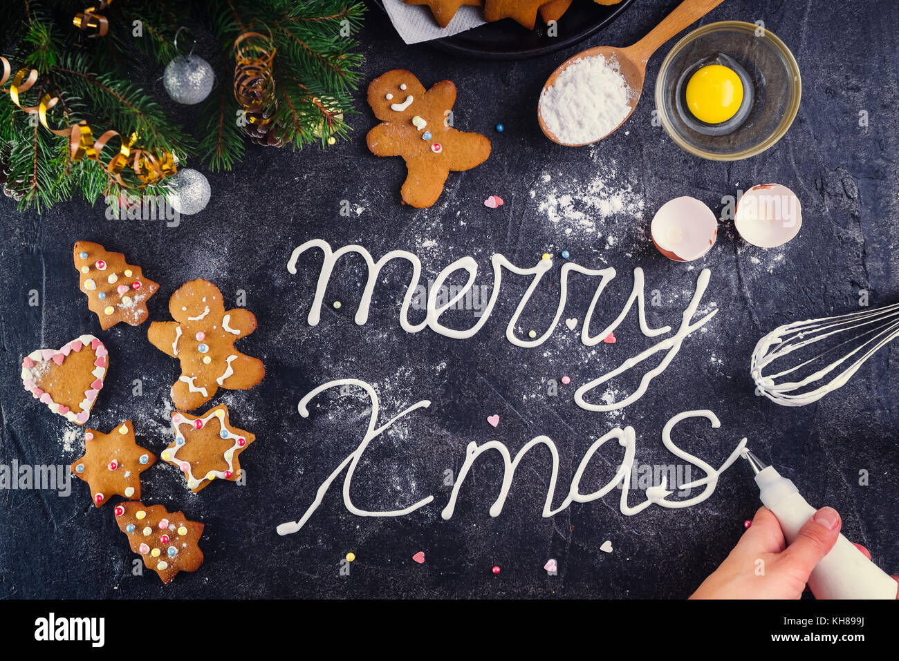 Vista superiore donna mani rendendo crema meringa scritte buon natale sulla pietra nera con sfondo ingrediens e dettagli decorativi. Anno nuovo concetto di vacanza. La cottura e il processo di decorazione. Foto Stock