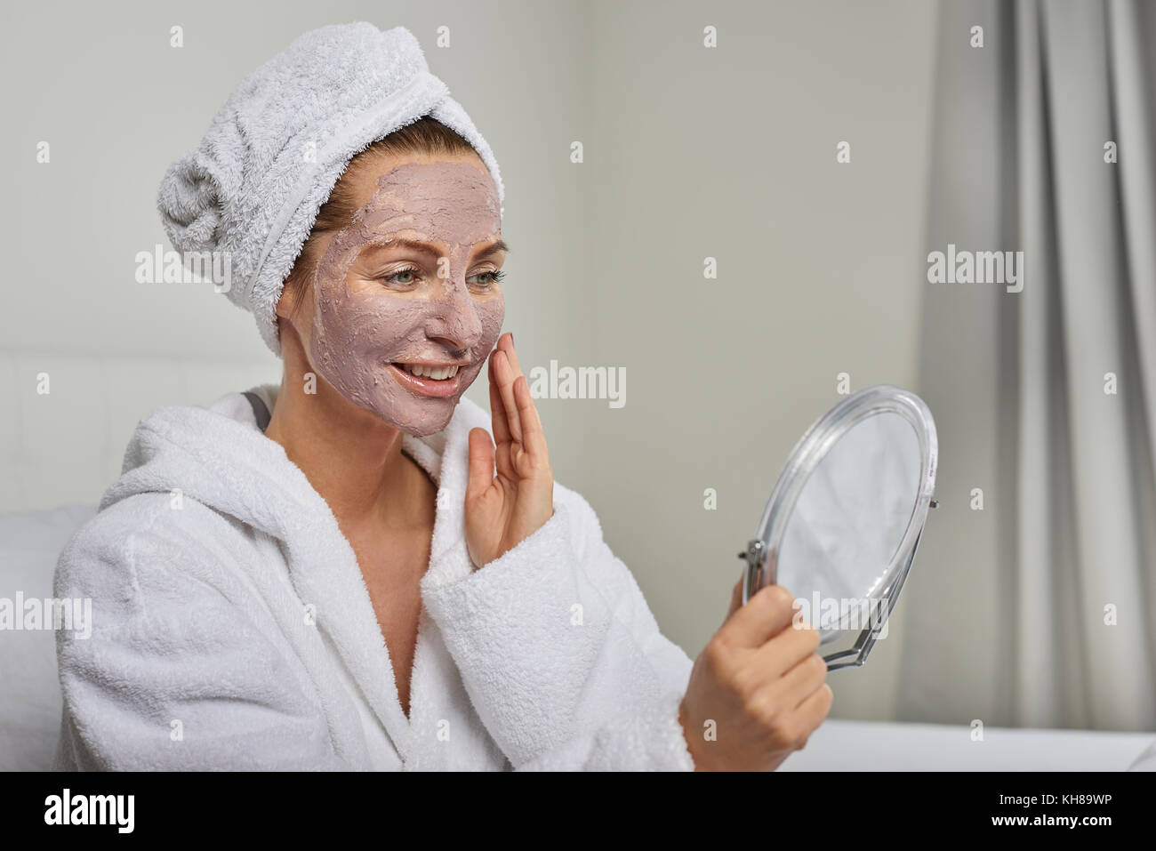 Donna attraente in un bianco accappatoio in spugna applicando una maschera per il viso mentre cercano in un palmare mirror come lei stessa vi vizierà con un trattamento di bellezza Foto Stock