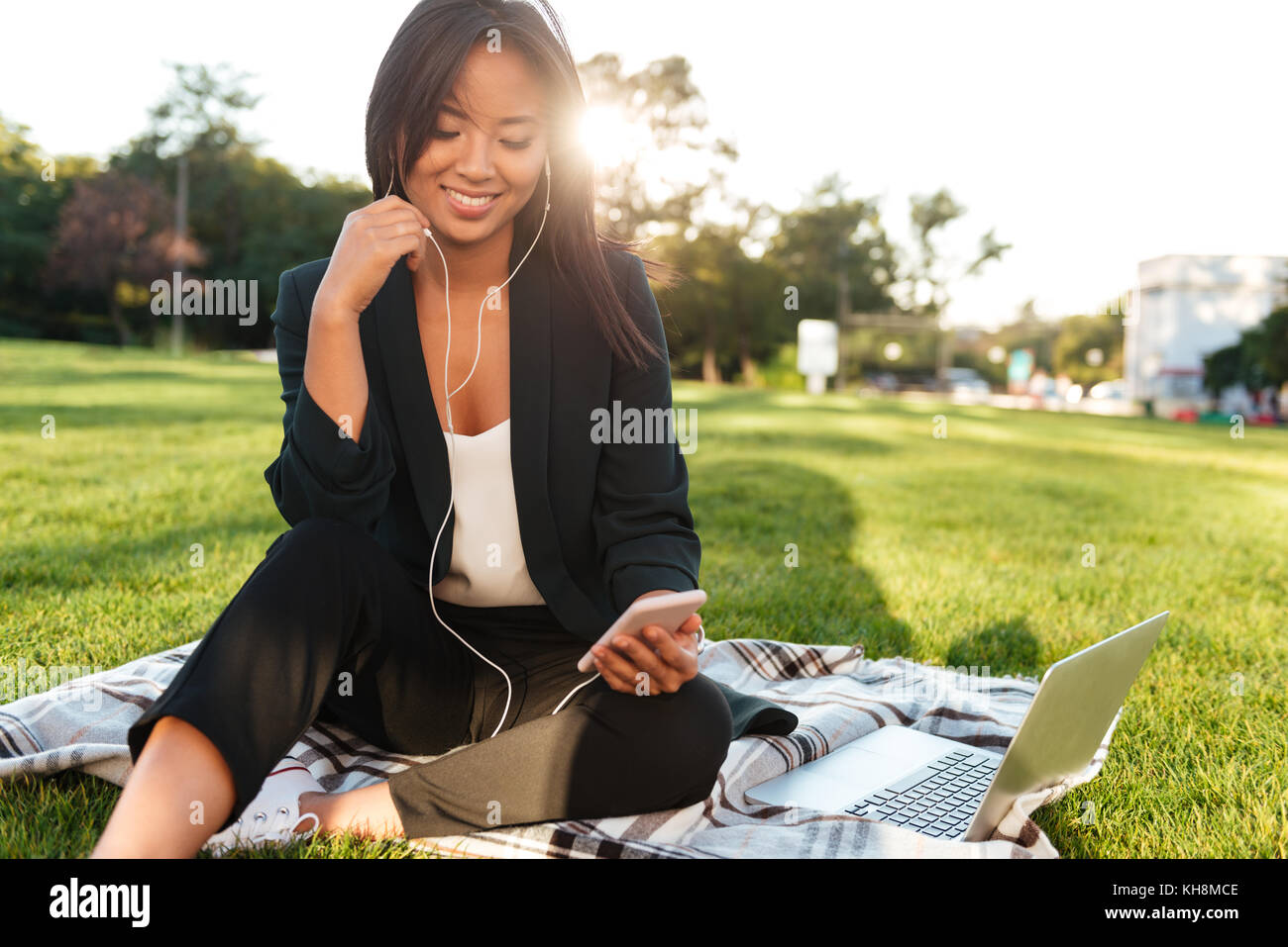 Pretty asian business donna, chattare su telefono cellulare, seduti su erba brillante Foto Stock