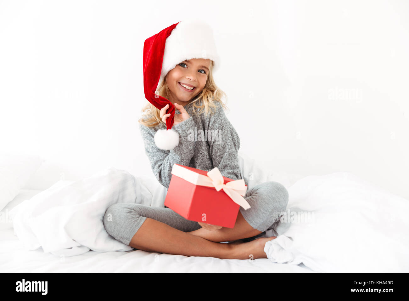 Affascinante piccolo ragazza di toccare la sua Santa's hat azienda confezione regalo, guardando la fotocamera mentre è seduto sul letto Foto Stock