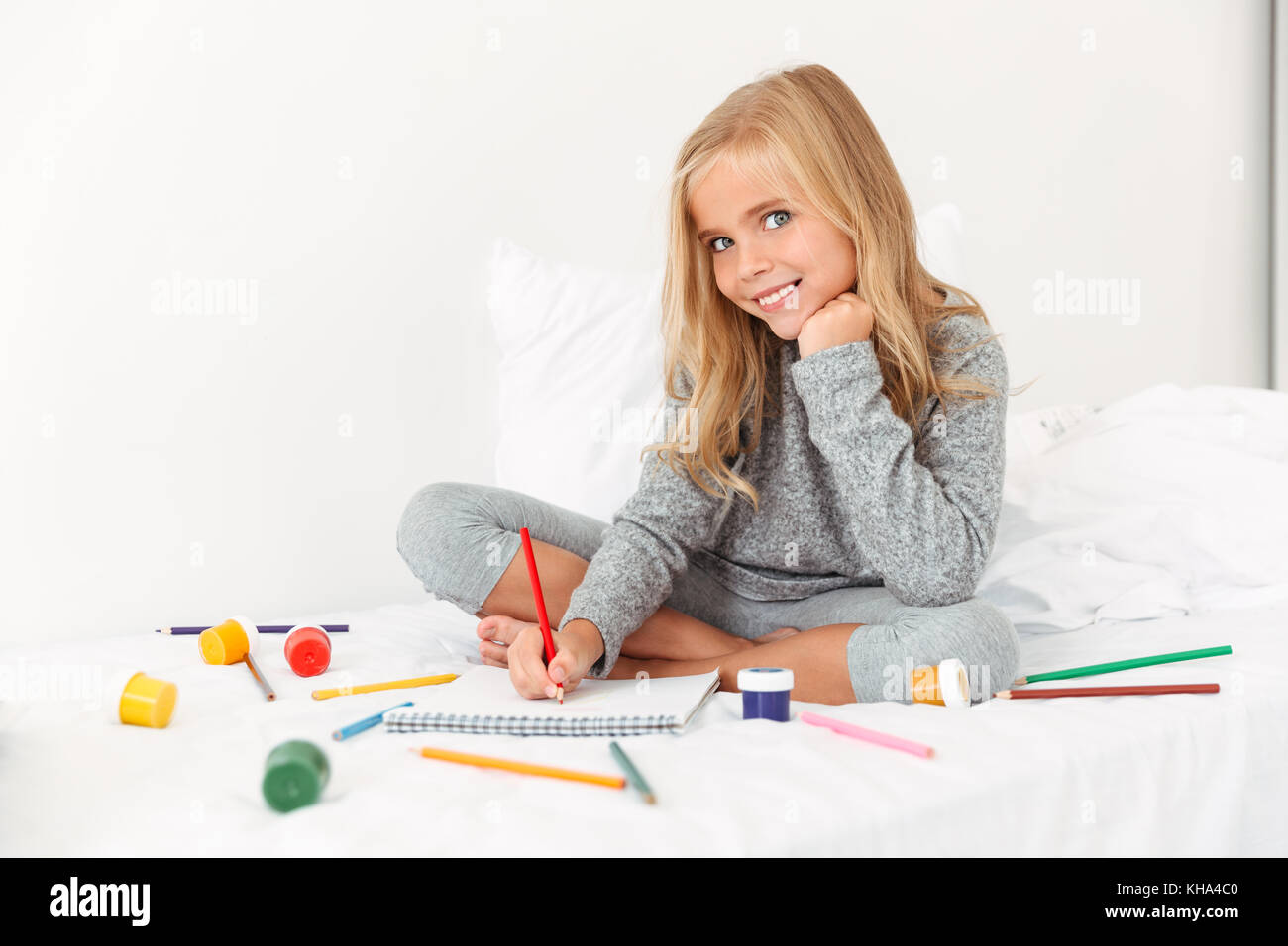 Affascinante piccolo ragazza Disegno con matite sul letto bianco, guardando la fotocamera Foto Stock