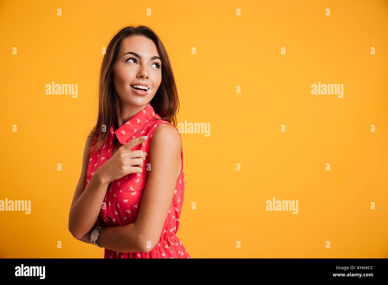 Ritratto di giovane stupito prety brunette woman in red dress puntando con il dito, cercando in disparte, isolato su sfondo giallo Foto Stock
