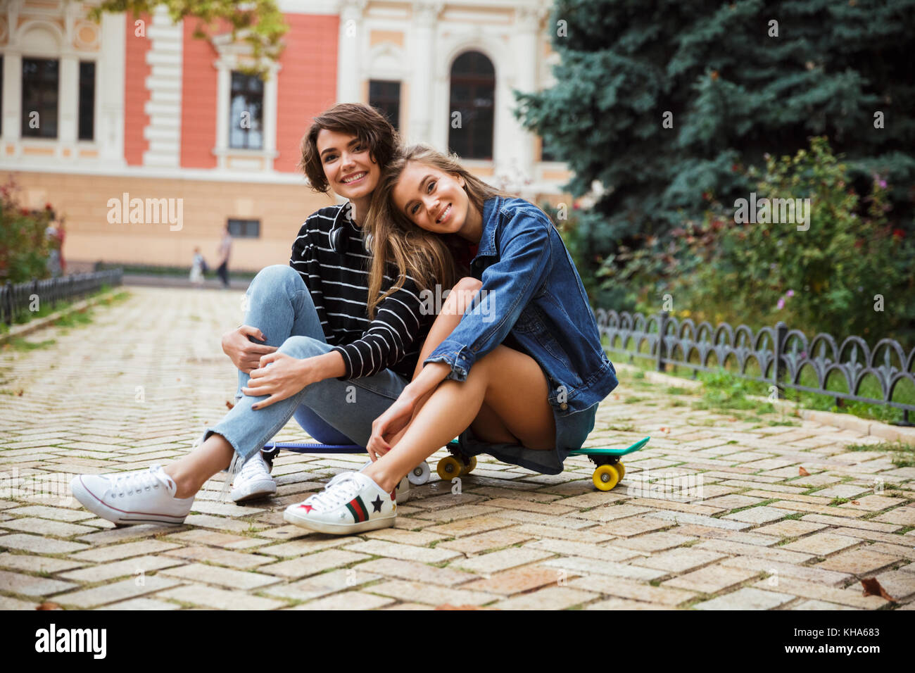 Due giovani sorridenti ragazze adolescenti seduti insieme su uno skateboard e guardando la telecamera su una strada di città all'aperto Foto Stock