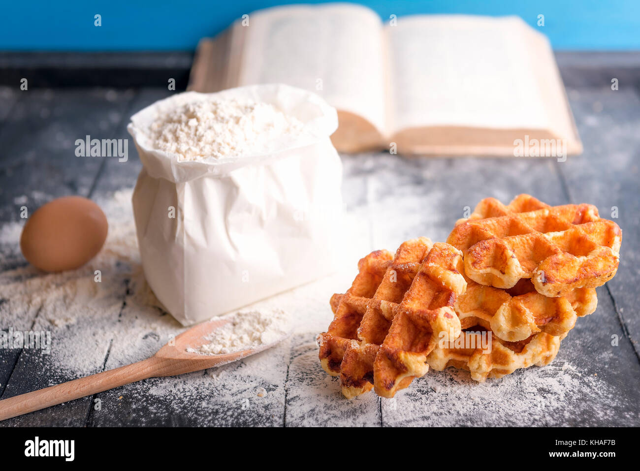 La cottura di immagine a tema con un mazzetto di deliziosi waffles belgi, uovo, un sacco di farina e un libro di cucina aperta, su una tavola in legno rustico. Foto Stock