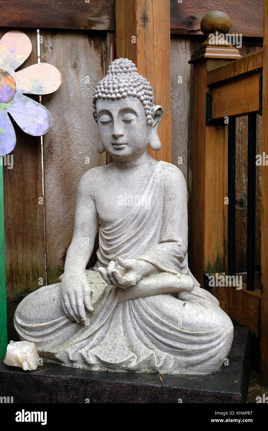 Serena Buddha statua di pietra in posizione seduta meditando pone sulla veranda in legno con fiore in background e cristallo in primo piano Foto Stock