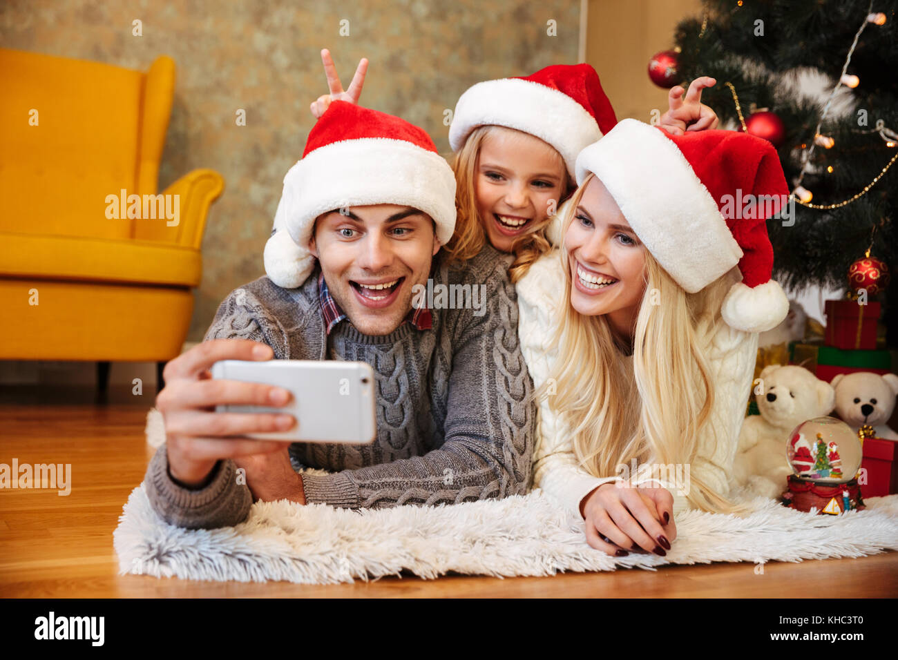 Felice bambina Santa's hat rende le corna ai suoi genitori mentre suo padre tenendo il Natale foto sul telefono cellulare Foto Stock