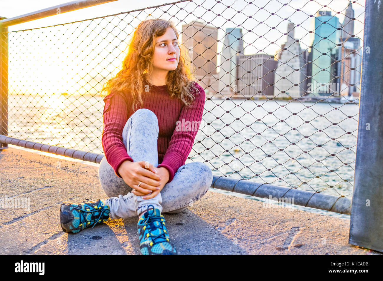 Primo piano di giovani hipster donna seduta dalla catena collegamento recinto nel ponte di Brooklyn Park si affaccia sul New York New York Manhattan cityscape skyline con acqua Foto Stock