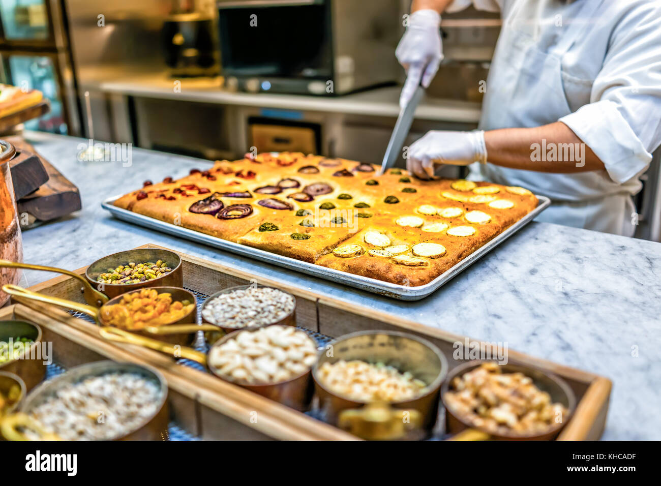 L uomo lo chef del ristorante cucina foccacia di taglio il pane con il coltello, noci, spezie in contenitori e condimenti dopo cottura in forno Foto Stock