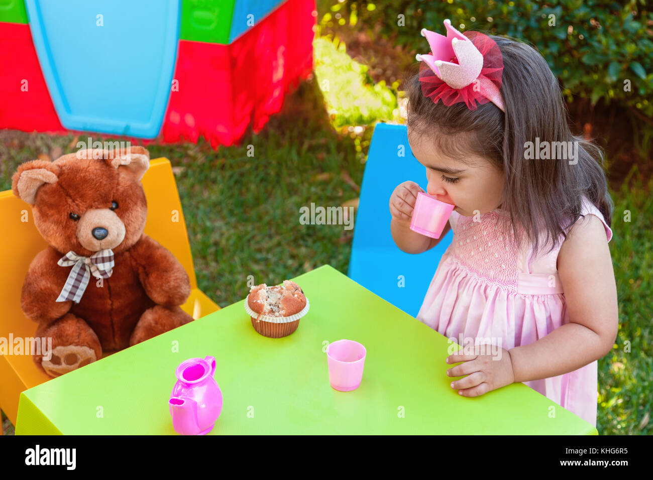 Baby toddler girl giocando in outdoor tea party di bere dal bicchiere con migliore amico orsacchiotto seduta a tavola. abito rosa e regina o princess crown Foto Stock