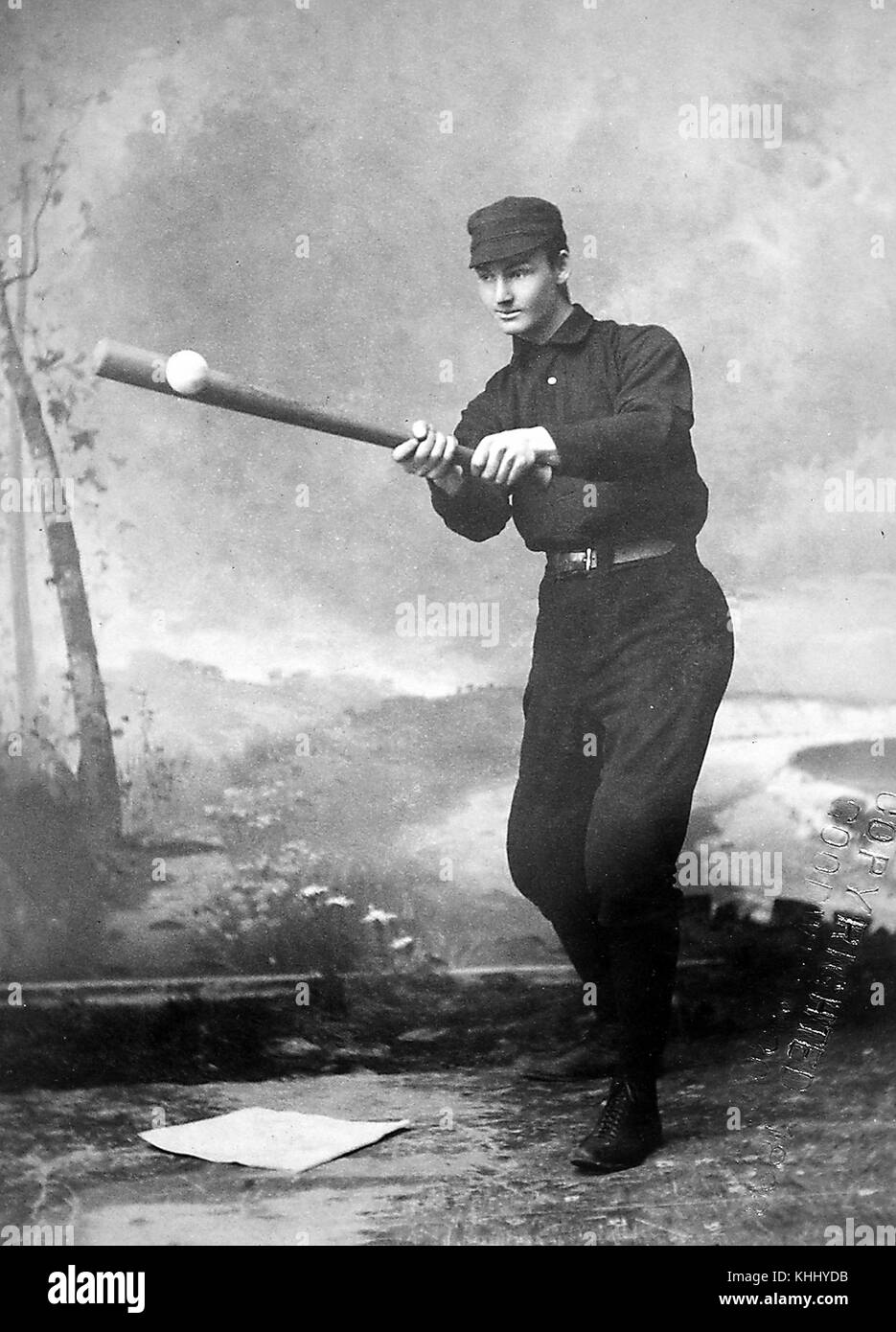 Un giocatore di baseball non identificato viene mostrato indossando un'uniforme scura, sta colpendo un baseball che gli è stato gettato da una persona che è fuori telaio, è posato davanti ad una scena paesaggistica dipinta che caratterizza alberi, erba, e un fiume, una base è stata posta sul pavimento di fronte a lui, 1900. Dalla Biblioteca pubblica di New York. Foto Stock